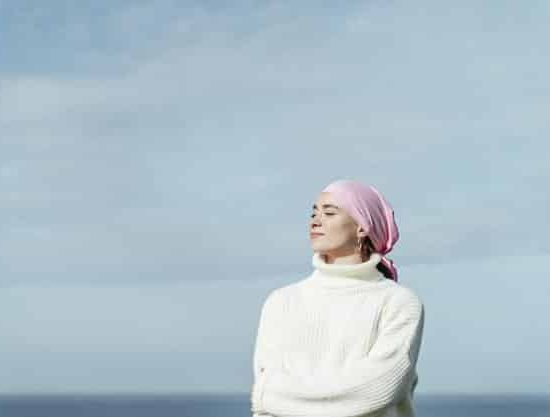 Young woman with cancer and crossed arms. She is with closed eyes. She is wearing a pink scarf on her head. She is feeling the sun on her face. She is wearing white jersey. Blue sky and sea on background